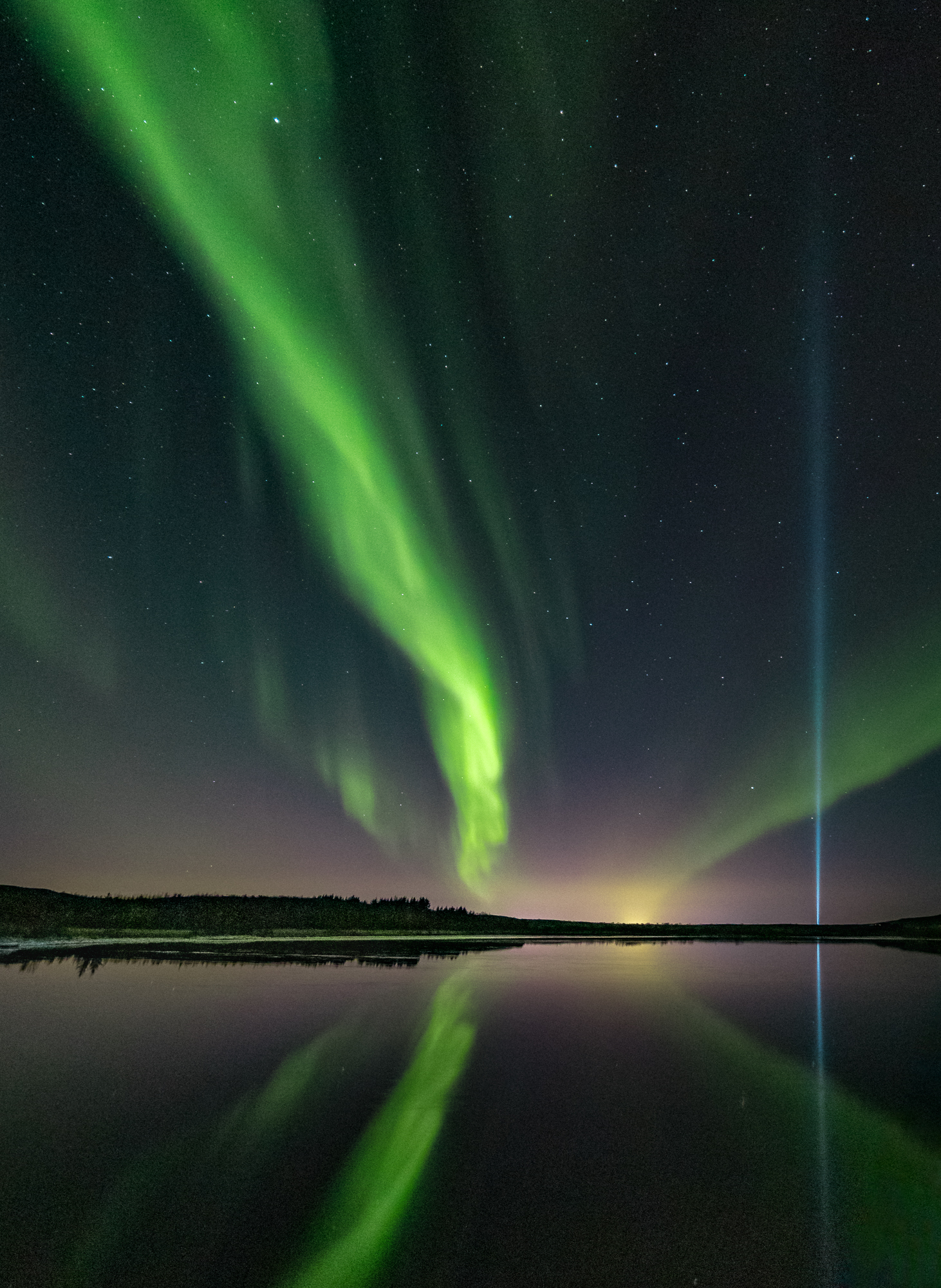 Natural and man-made lights brighten the night sky in Iceland. The blue pillar is the Imagine Peace Tower (Friðarsúlan) light installation by Yoko Ono (a memory for John Lennon). Reykjavik lights shimmer on the horizon. These modern lights compete in brilliance with stars and auroras of the “old world”.
I took the photo on my trip to Iceland in late 2019. On the very last night of my stay there was a vivid aurora storm, so I drove to the outskirts of Reykjavik to watch and photograph it. The light of Friðarsúlan added a special twist to this magnificent display.